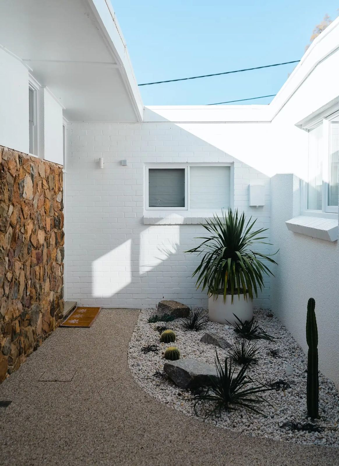 White-brick courtyard with a random-rubble stone wall, white-pebble desert bed, barrel cacti, dracaena and rocks