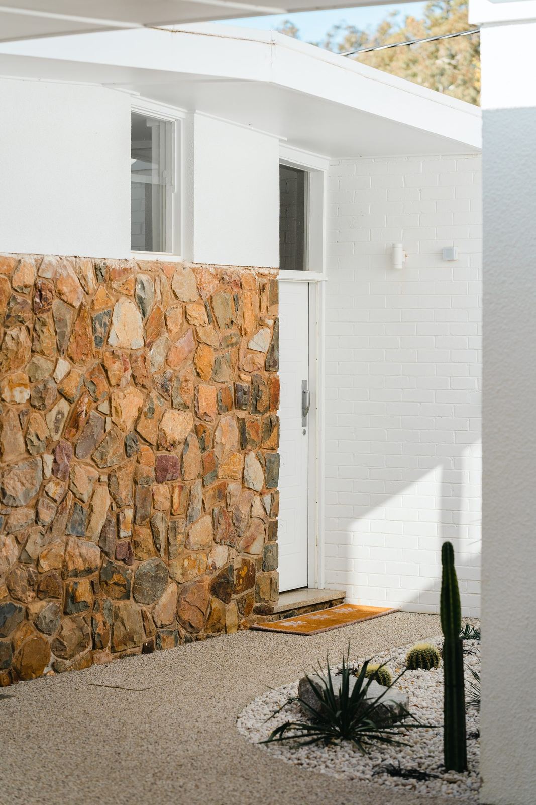 Random-rubble stone feature wall beside the white front door, with exposed-aggregate paving and cacti