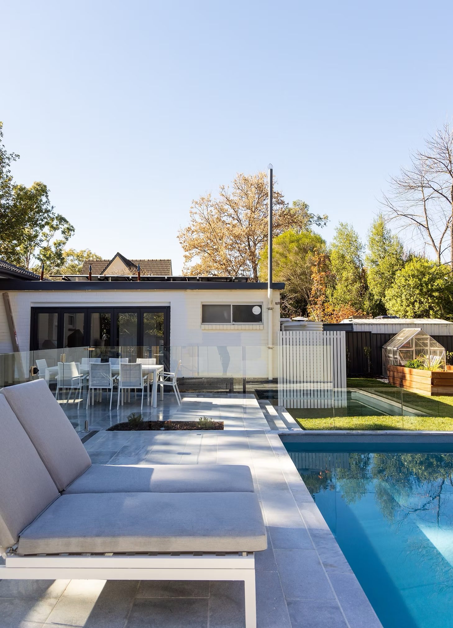 Built-in outdoor lounge with cushions beside the pool, house and greenhouse beyond