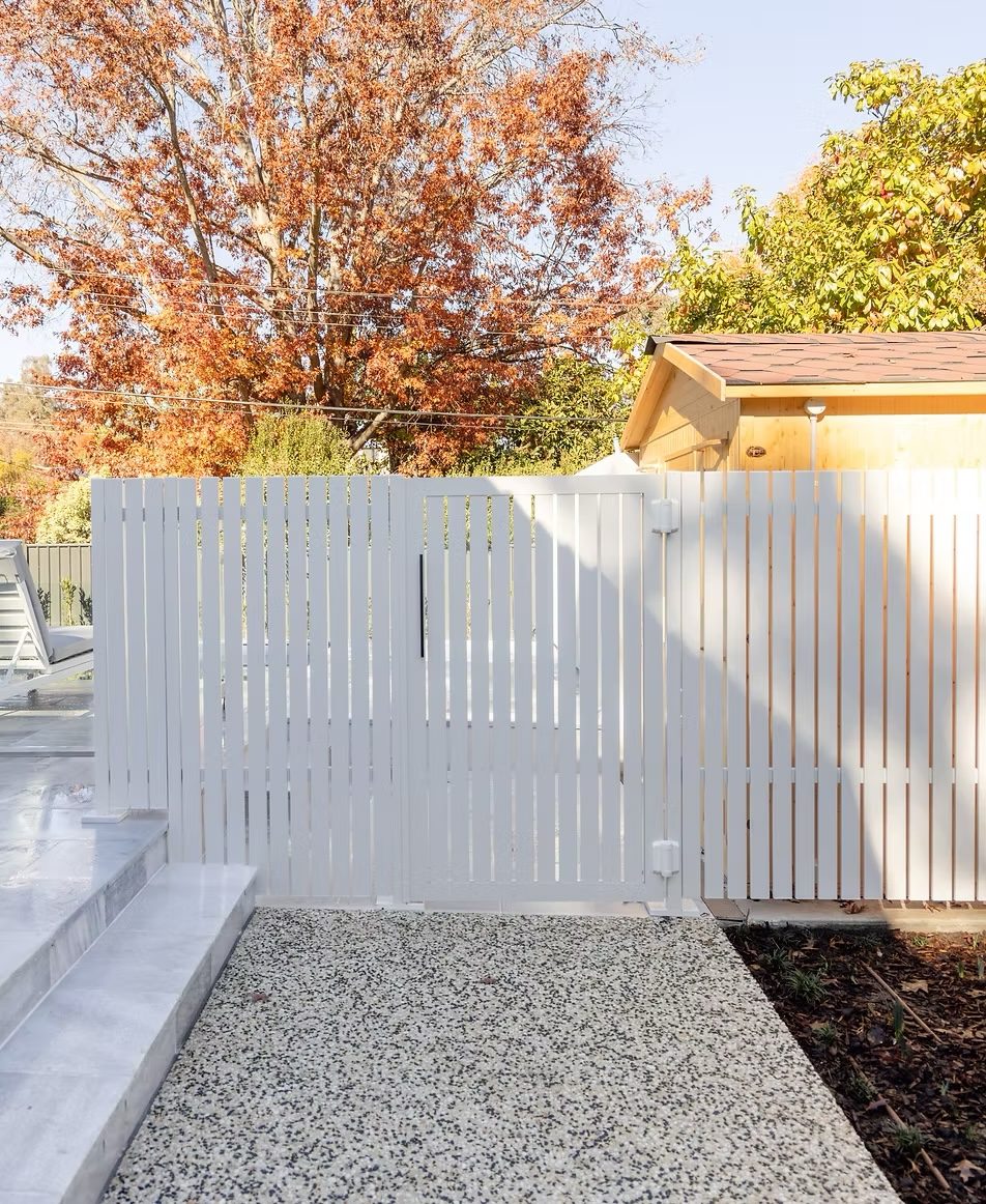 White vertical-batten pool gate on gravel with an autumn tree overhead