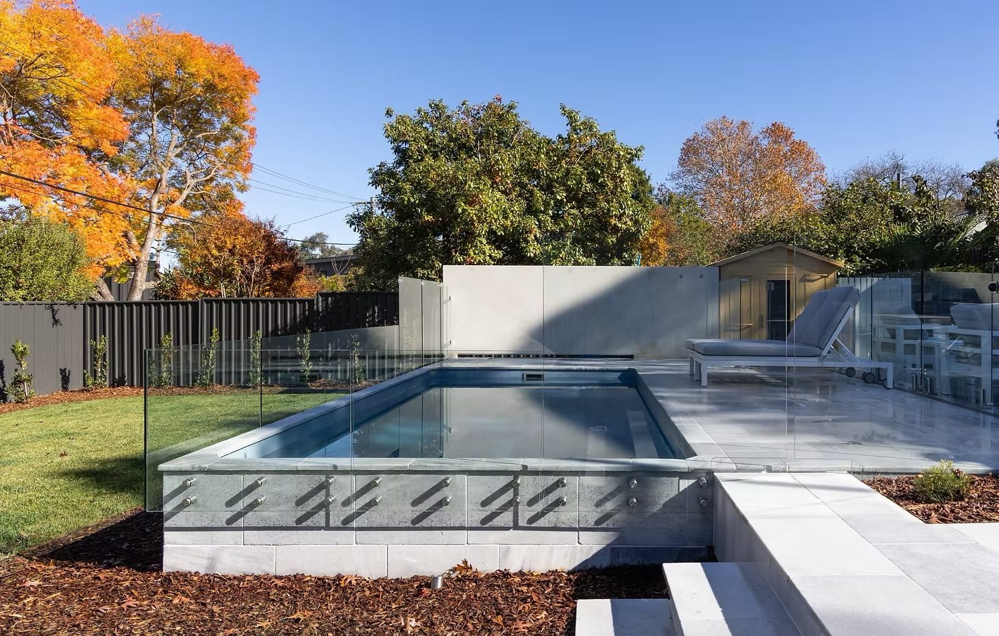 Garran: backyard pool and spa with a glass fence, rendered wall and autumn garden