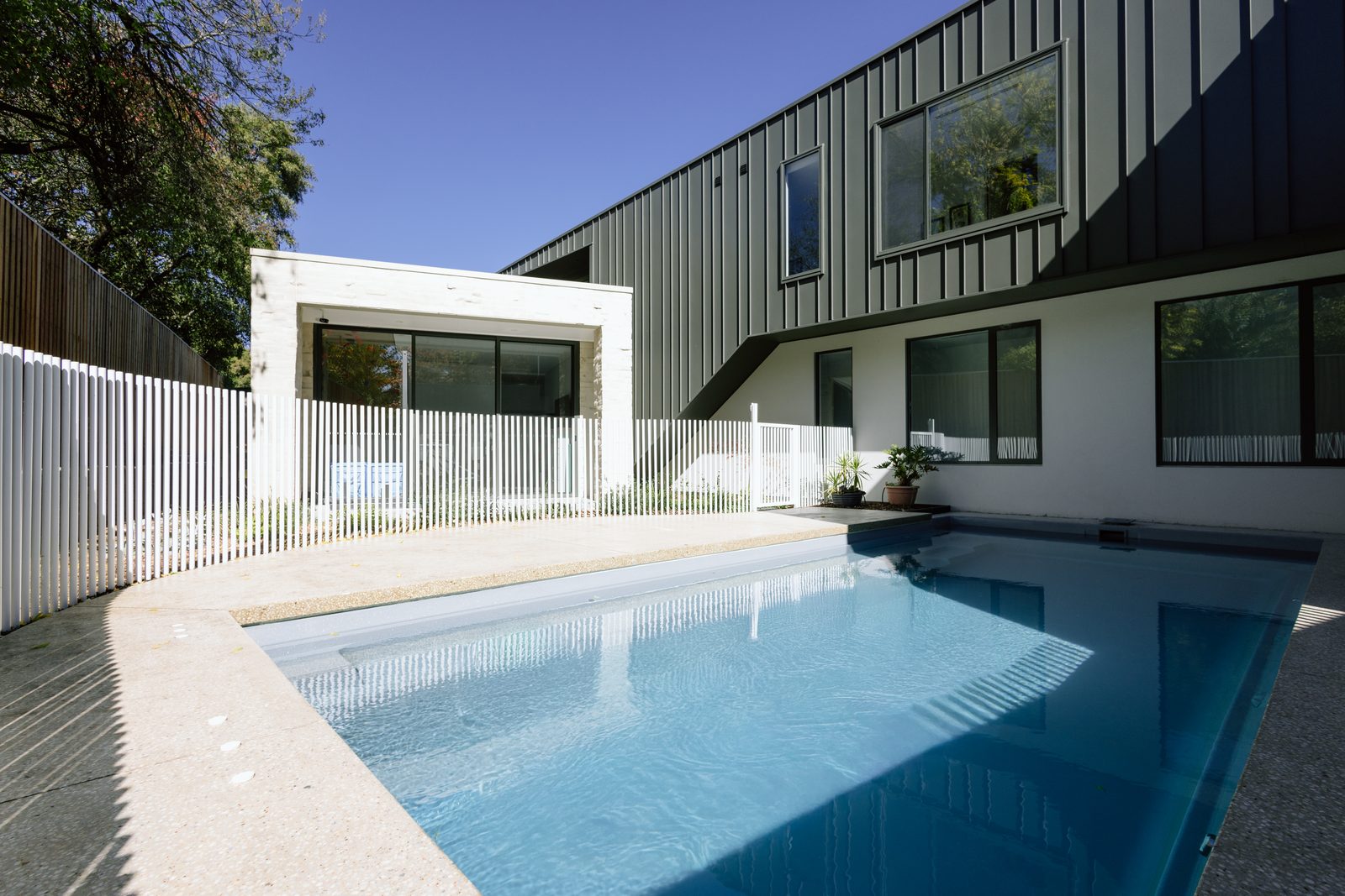 Turquoise lap pool with a white picket fence beside the dark standing-seam home