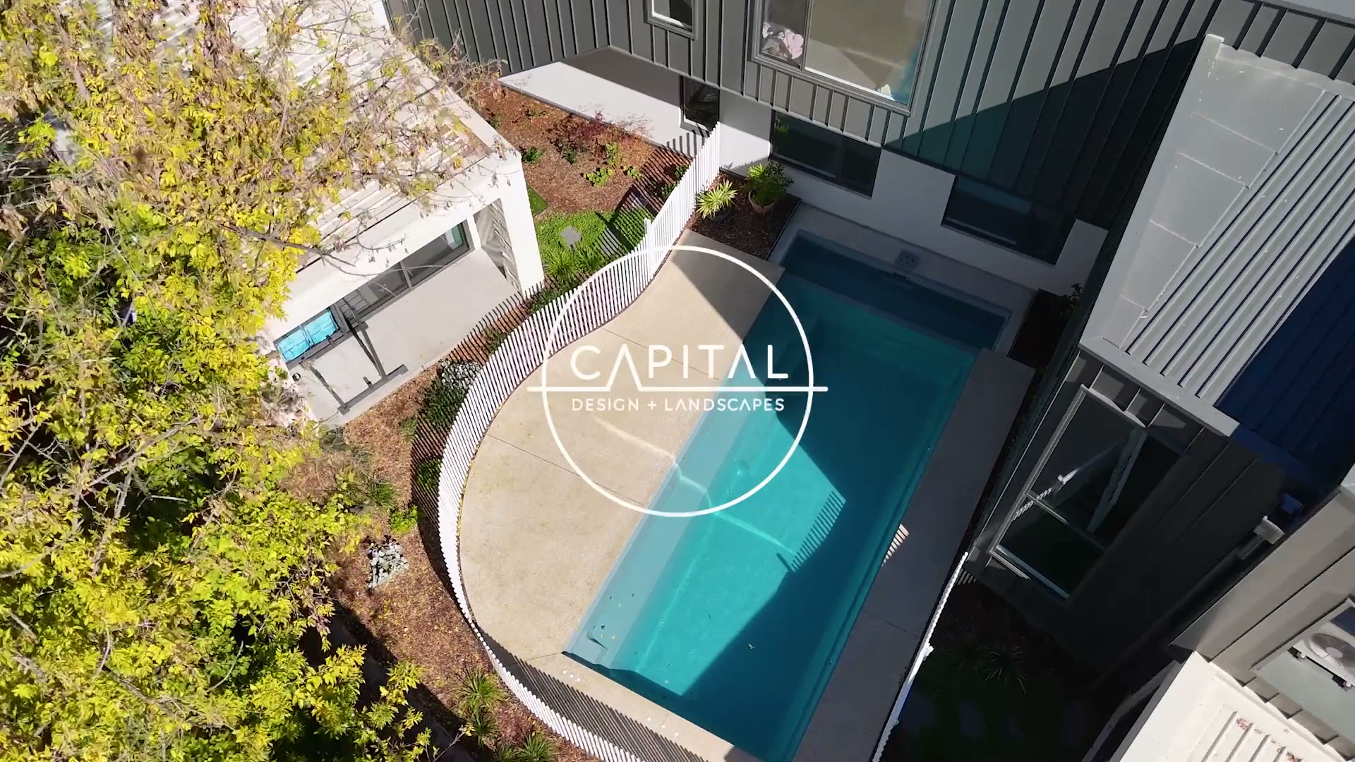 Finished Yarralumla pool from above: turquoise water, exposed-aggregate surround and curved timber picket fence
